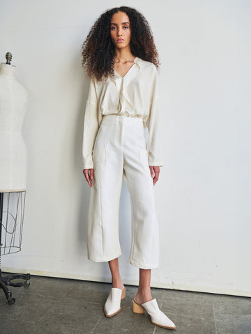 In a minimalist, fashion-focused setting emphasizing sustainable materials like organic cotton, an individual with curly hair stands against a plain wall. They are adorned in a light-colored blouse and the Curved Eda Culotte by Zero + Maria Cornejo, paired with white shoes. A mannequin is partially visible on the left.