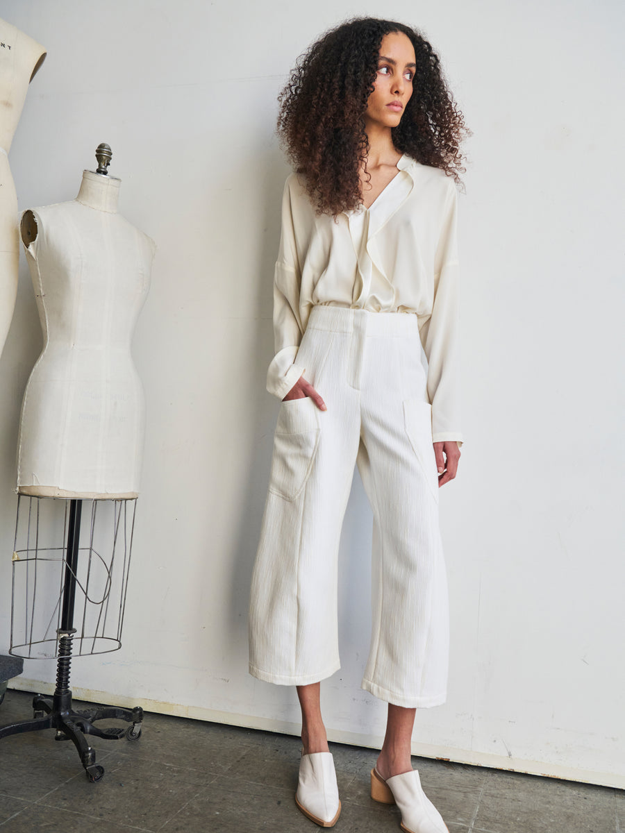 In a minimalist room, a person with curly hair stands confidently next to mannequins, exuding style as they wear the Curved Eda Culotte from Zero + Maria Cornejo paired with a white blouse, their hands casually tucked into their pockets.