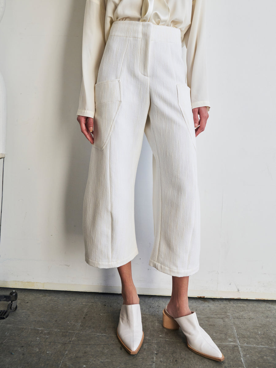 The individual is dressed in the Curved Eda Culotte by Zero + Maria Cornejo, crafted from organic cotton in a cream hue and adorned with large pockets, complemented by a matching blouse. They have styled the outfit with white heeled mules, standing against a plain white wall and concrete floor backdrop.