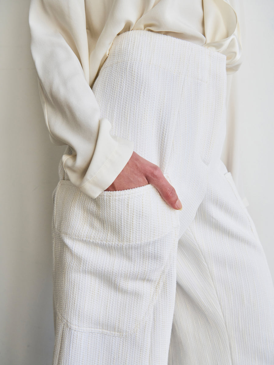 Dressed in the Curved Eda Culotte by Zero + Maria Cornejo, an individual is seen wearing a textured, white outfit made from organic cotton. The ensemble looks both comfortable and stylish, featuring a long-sleeved top and high-waisted pants with a loose fit, with their hand casually tucked into a pocket.