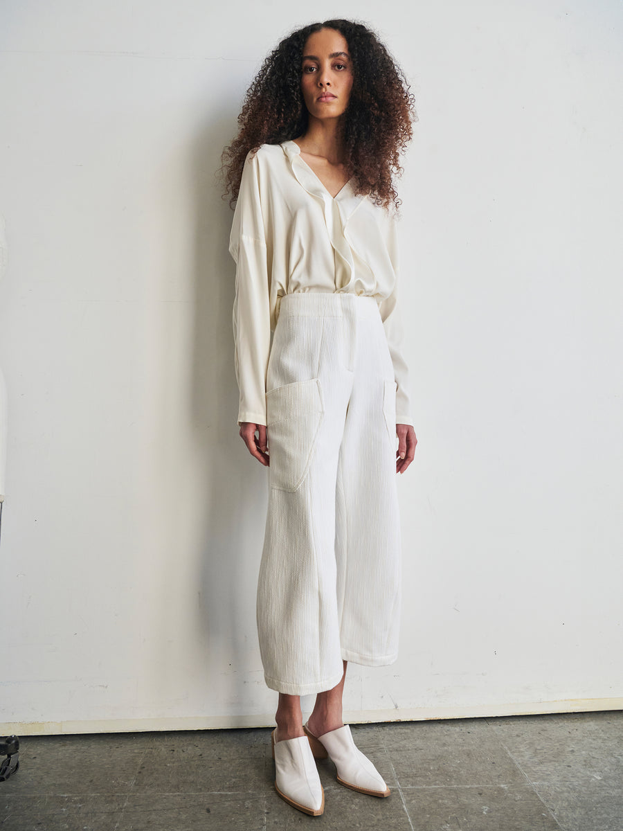 A person with curly hair is wearing a white blouse and Curved Eda Culotte from Zero + Maria Cornejo, paired with white shoes. They are standing against a plain background, exuding a relaxed and stylish vibe.