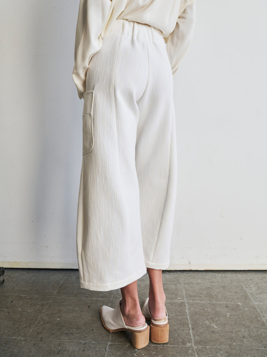 A person is seen from behind wearing an outfit crafted from organic cotton, showcasing the Curved Eda Culotte by Zero + Maria Cornejo. The ensemble includes a cream-colored long-sleeved top and wide-legged pants with large side pockets, perfectly complemented by slip-on block heels. A gray floor and a plain light wall serve as a minimalist backdrop.