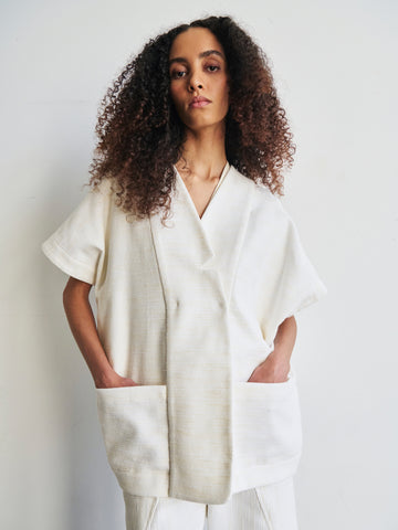 A person with long curly hair is wearing the Saban Waistcoat, an oversized white kimono-style top from Zero + Maria Cornejo made from organic cotton, featuring short sleeves. Their hands rest in the large front pockets as they stand against a plain light-colored background.