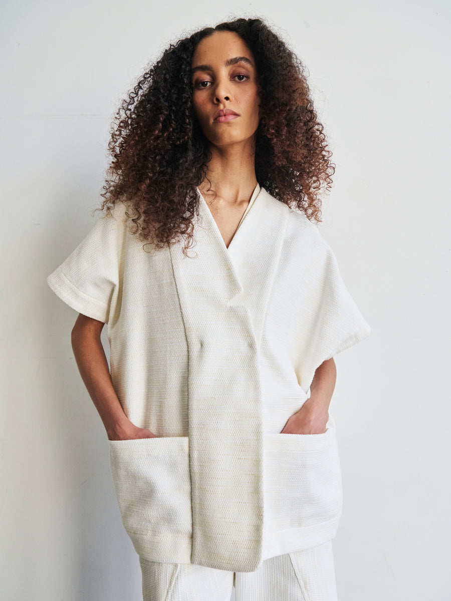 A person with long curly hair is wearing the Saban Waistcoat, an oversized white kimono-style top from Zero + Maria Cornejo made from organic cotton, featuring short sleeves. Their hands rest in the large front pockets as they stand against a plain light-colored background.