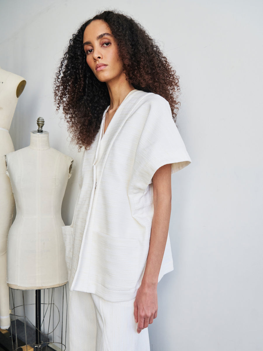 A person with curly hair wearing the Saban Waistcoat by Zero + Maria Cornejo, made from organic cotton, stands beside dress forms. The outfit includes a short-sleeved, loose-fitting top and matching pants. The neutral background enhances the minimalist setting.