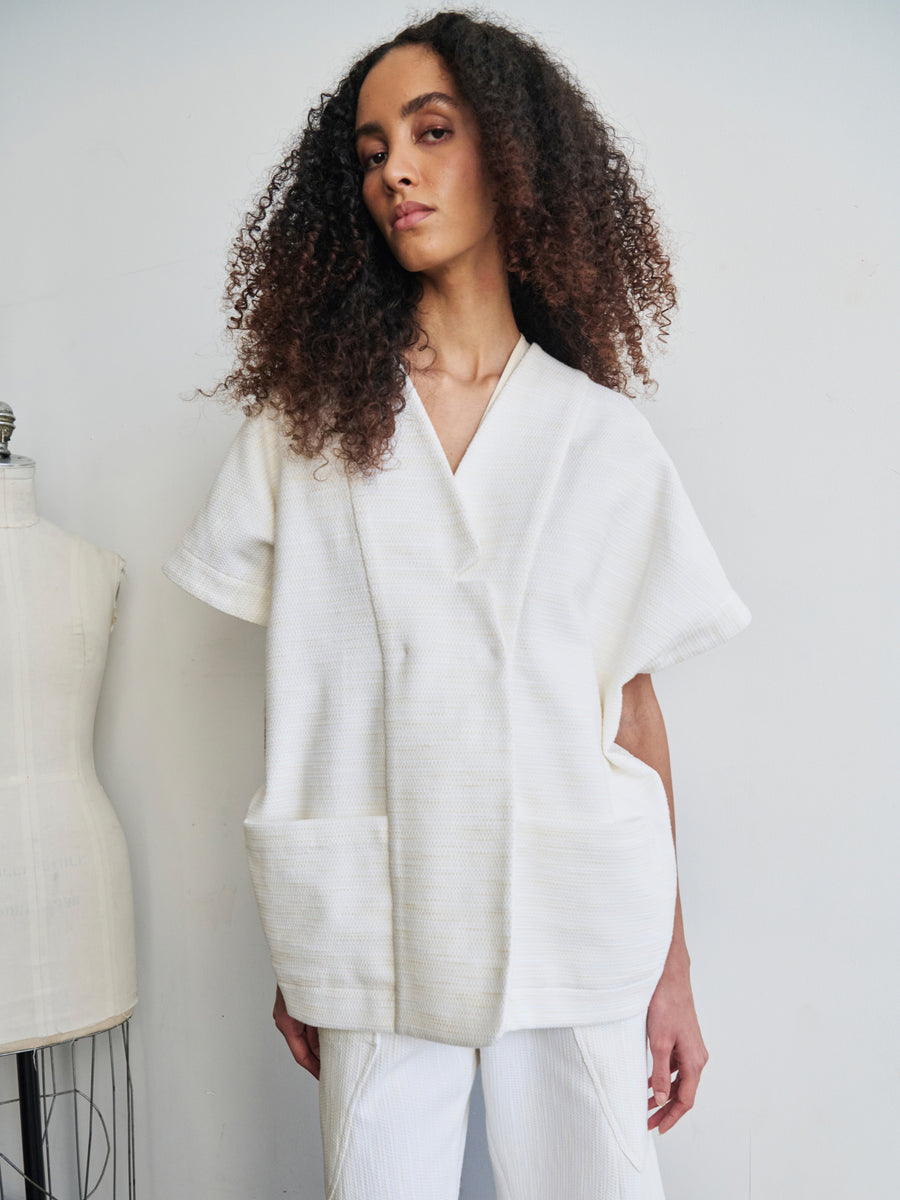 A person with curly hair stands against a plain background, dressed in the Saban Waistcoat by Zero + Maria Cornejo. The loose-fitting, white top is crafted from organic cotton and features large pockets, which is paired with matching white pants. A dress form is partially visible on the left.