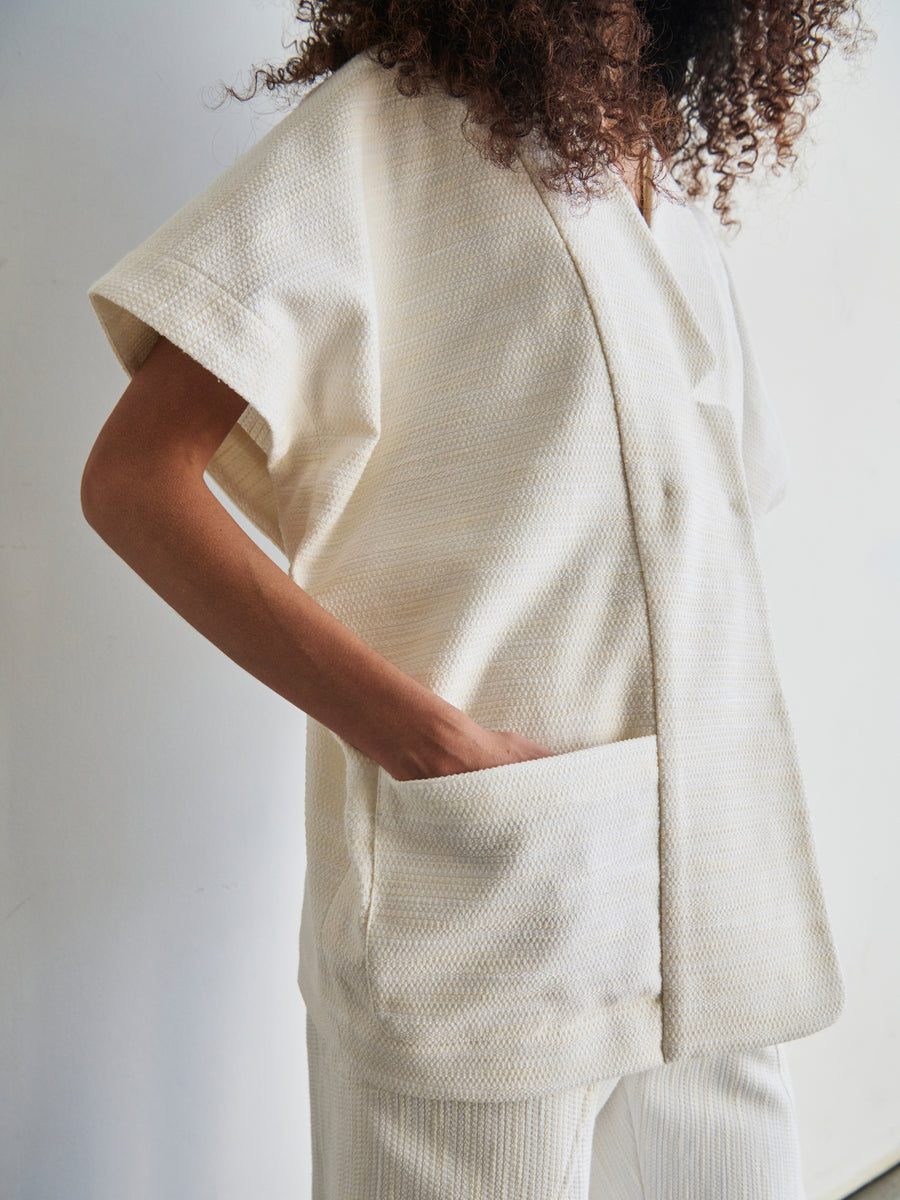 A person with curly hair models the Saban Waistcoat by Zero + Maria Cornejo, a cream-colored, textured outfit crafted from organic cotton. It features a loose-fitting top with short sleeves and a pocket, complemented by matching pants. The setting is minimalist with a soft, neutral background.