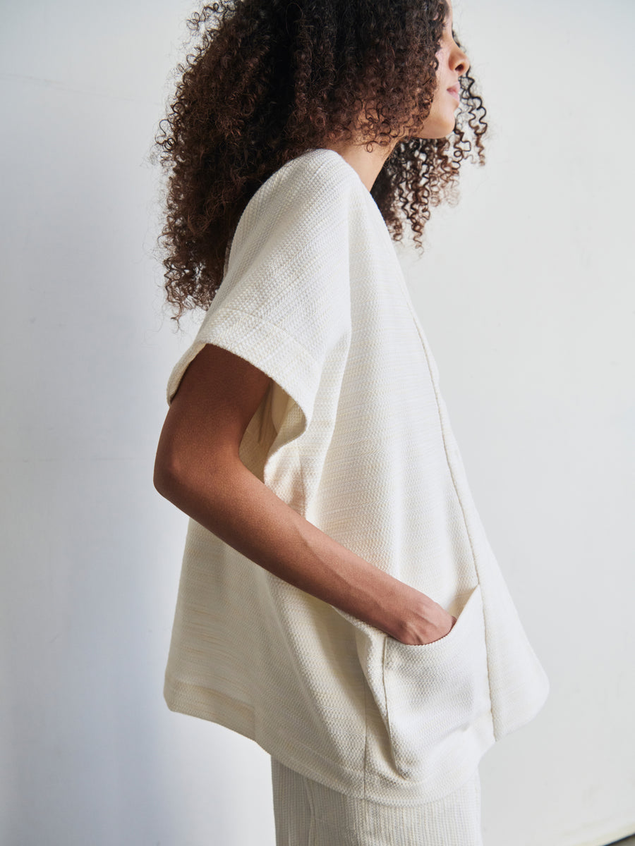 A person with curly hair is standing sideways, wearing the Saban Waistcoat by Zero + Maria Cornejo, a textured white short-sleeve top made from organic cotton and featuring pockets. The scene is set against a plain background.