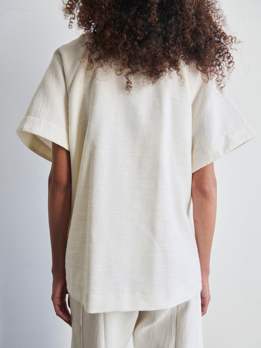 Rear view of a person with curly hair wearing the Saban Waistcoat by Zero + Maria Cornejo, featuring a loose-fitting, short-sleeved cream-colored top paired with matching pants made from recycled polyester. The textured fabric enhances the minimalist vibe as the individual stands against a plain white background.