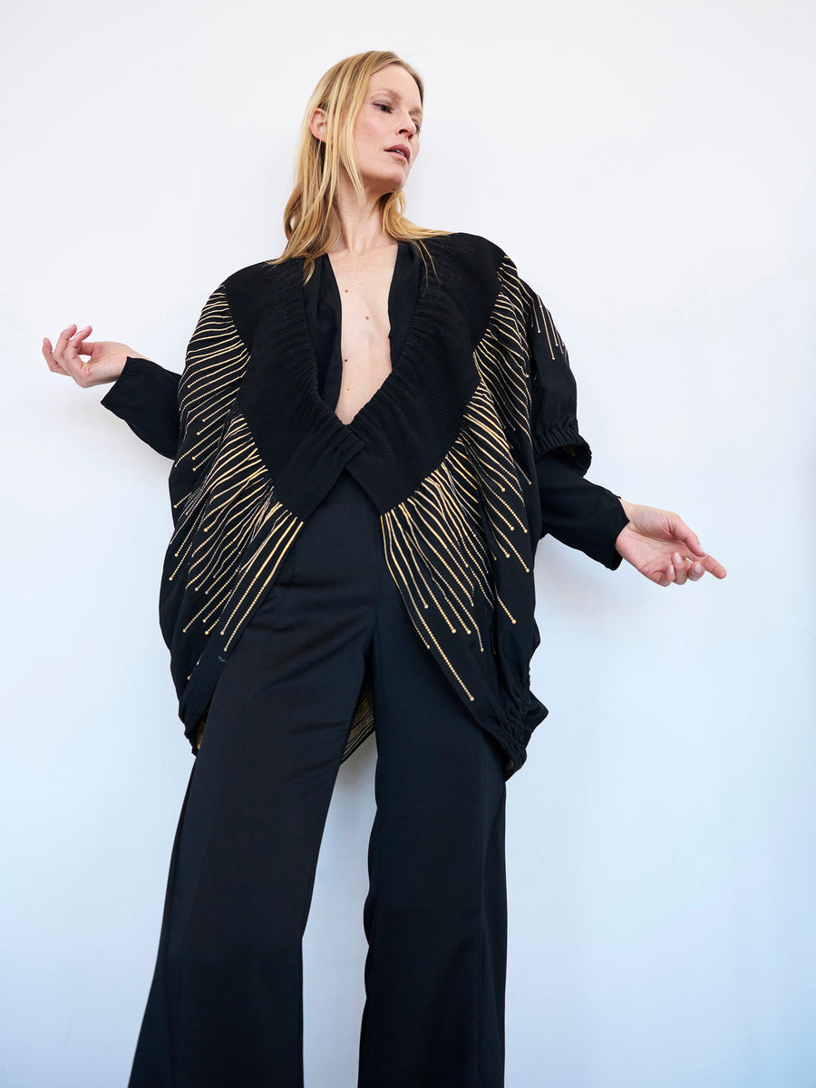 A woman with long blonde hair poses against a plain white background. She is wearing a black wide-legged jumpsuit with a deep v-neckline and the Evia Shrug by Zero + Maria Cornejo, adorned with organic metallic smocking. Her arms are extended outwards, and she gazes off to the side.