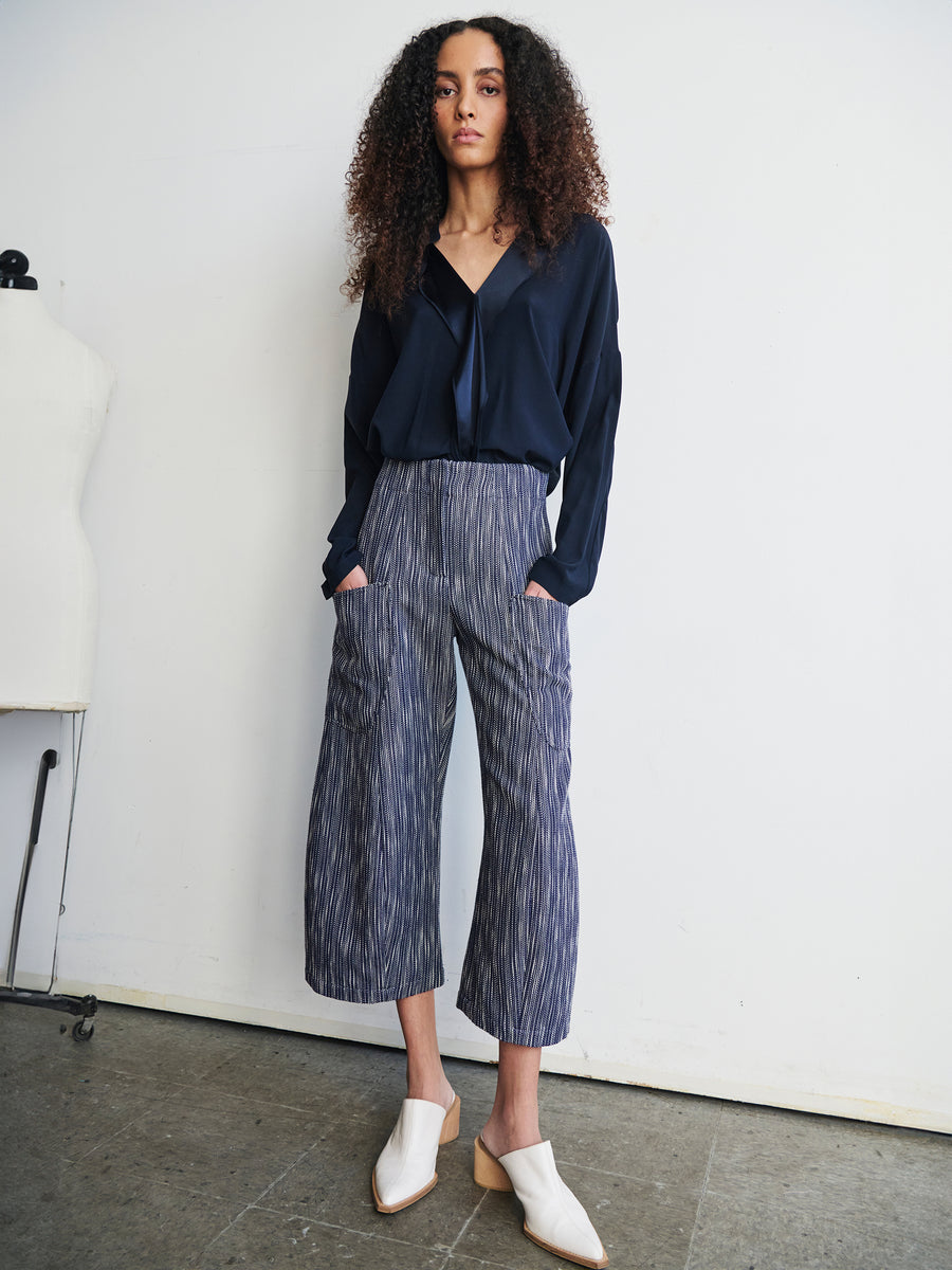 Against a white wall stands a person with curly hair, dressed in a navy blouse and the Curved Eda Culotte by Zero + Maria Cornejo, made from organic cotton. Their hands rest in the pockets of their striped wide-legged pants, and they finish the look with white shoes.