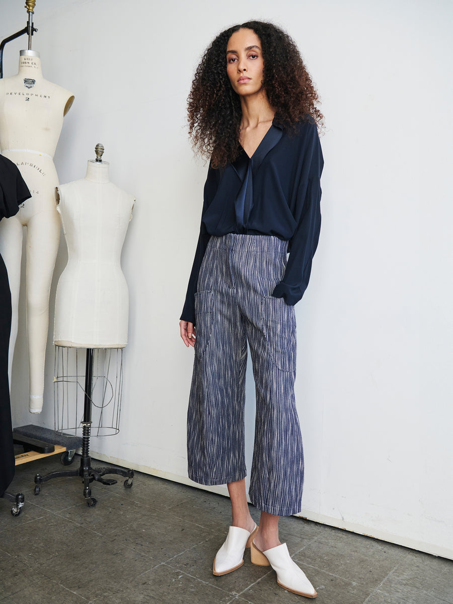 A person with long curly hair is dressed in a dark blouse and the Curved Eda Culotte by Zero + Maria Cornejo, made from organic cotton tweed. They are surrounded by dressmaker mannequins while wearing white slip-on shoes and standing on a textured gray floor.