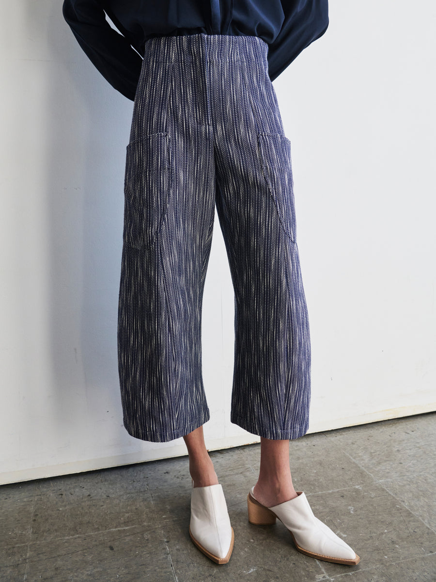 Someone wearing the Curved Eda Culotte by Zero + Maria Cornejo, featuring blue-and-white stripes in organic cotton, stands against a white wall. They pair it with white slip-on block heels and a navy blue top while posing with their hands behind their back on a floor that displays a grey stone texture.