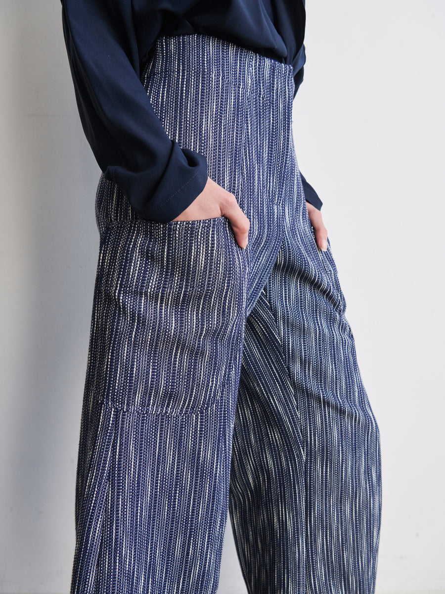 A person wearing a dark long-sleeve shirt and the Curved Eda Culotte by Zero + Maria Cornejo, designed with high-waisted blue and white stripes and crafted from recycled polyester, stands with their hands in the pockets against a plain white background.