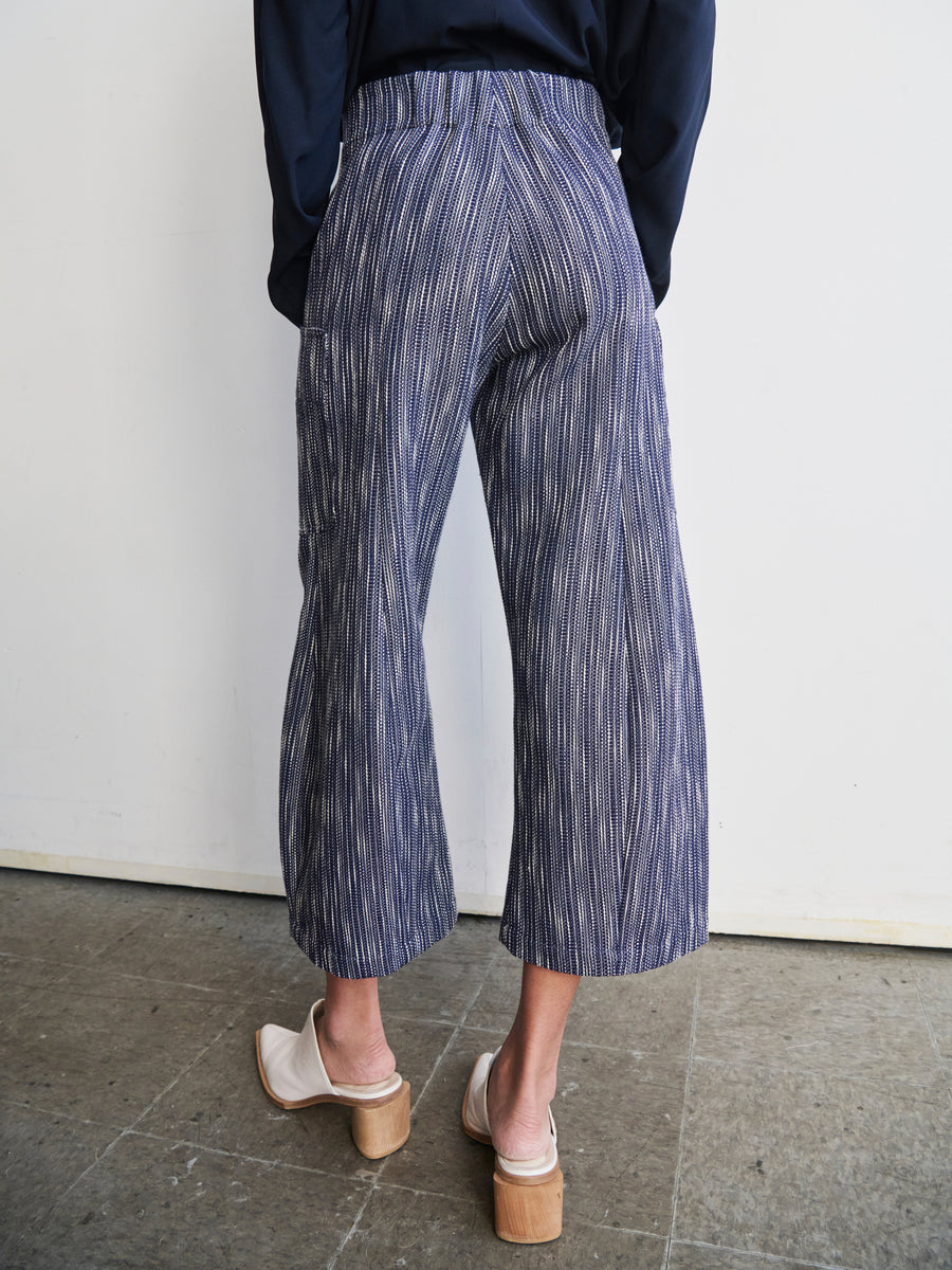 A person wearing the Curved Eda Culotte by Zero + Maria Cornejo, featuring blue and white stripes and made from organic cotton, pairs the look with white heeled clogs as they stand on a stone floor facing a plain white wall.