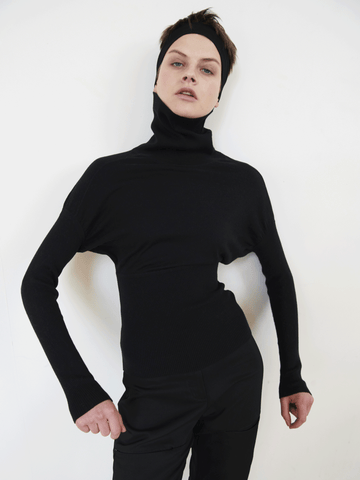 A person with short hair is wearing a sleek black Long-Sleeved Ama Rollneck by Zero + Maria Cornejo made from breathable luxury fabric and a matching black headband. They stand against a plain white background, with one hand resting on their hip and the other hanging by their side, striking a confident pose.