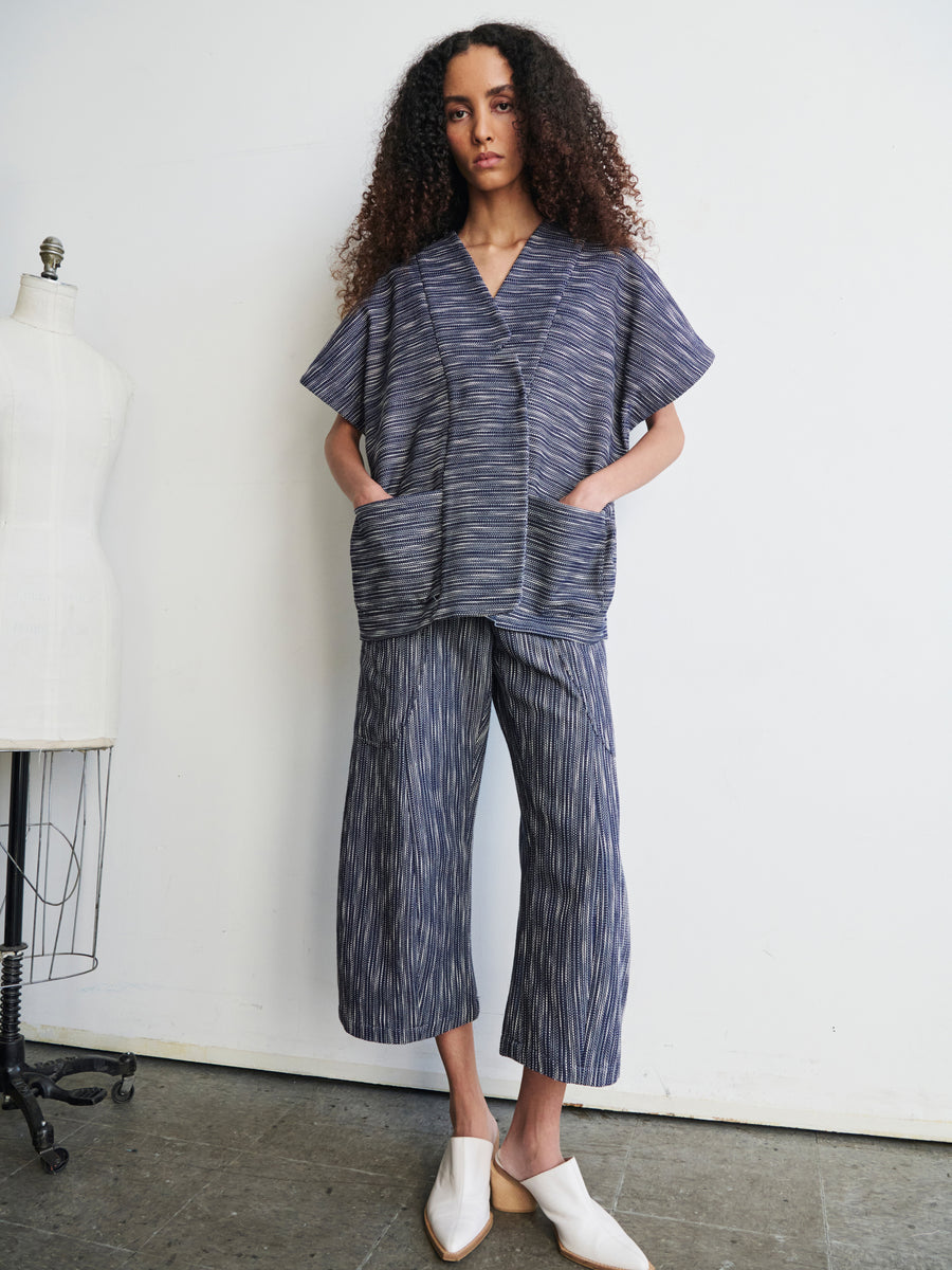 A person with curly hair is wearing the Saban Waistcoat, a loose-fitting striped piece made of organic cotton from Zero + Maria Cornejo. They stand near a dress form, hands in pockets, accompanied by white shoes against a plain and minimalist background.