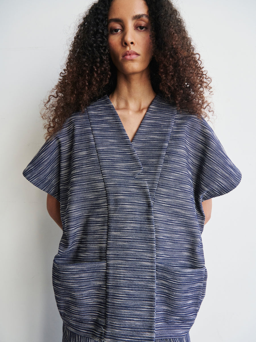 A person with long, curly hair is wearing the Saban Waistcoat by Zero + Maria Cornejo, a textured, short-sleeved garment in blue-gray organic cotton. They stand against a plain, light background, looking directly at the camera.