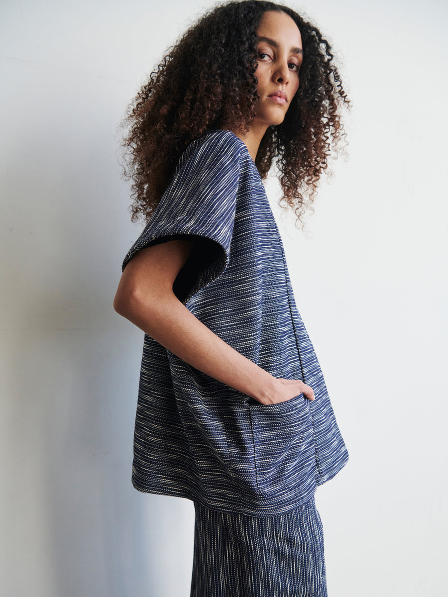 A person with curly hair is wearing the Saban Waistcoat by Zero + Maria Cornejo, a loose-fitting, textured garment crafted from organic cotton in dark blue hues. They stand against a plain background, gazing to the left, with hands casually tucked in pockets.