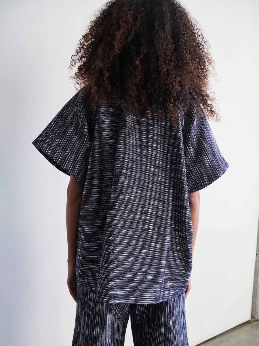 A person with long curly hair is wearing a dark blue Saban Waistcoat with a textured finish, created by Zero + Maria Cornejo, and matching pants, all crafted from organic cotton. They stand facing away from the camera against a white background.