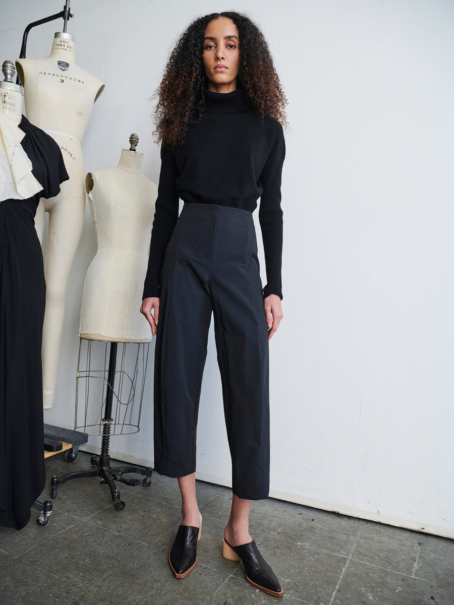 A person with long curly hair stands confidently in a fashion design studio, wearing a black turtleneck and the Margot Pant from Zero + Maria Cornejo. Their ensemble is completed by black loafers. In the background, mannequins dressed in various garments enhance the creative ambiance of the pristine white-walled studio.