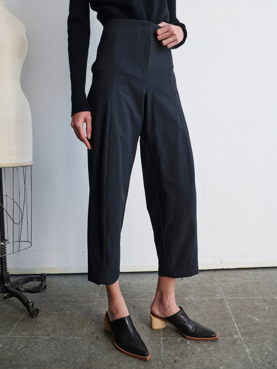 A person stands wearing high-waisted black Margot Pants from Zero + Maria Cornejo and black mules with wooden heels. The outfit is completed with a black long-sleeved top. Part of a dress form is visible on the left side, standing on a tiled floor against a white wall.