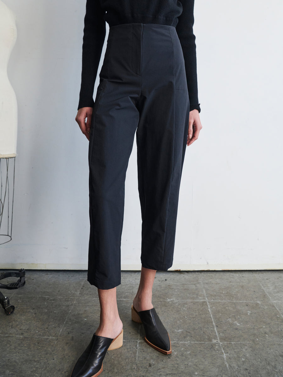 A person stands in a minimalist room with gray flooring, wearing black Margot Pants by Zero + Maria Cornejo and a black sweater. Black mules complete the sleek look, with part of a dress form visible in the background.