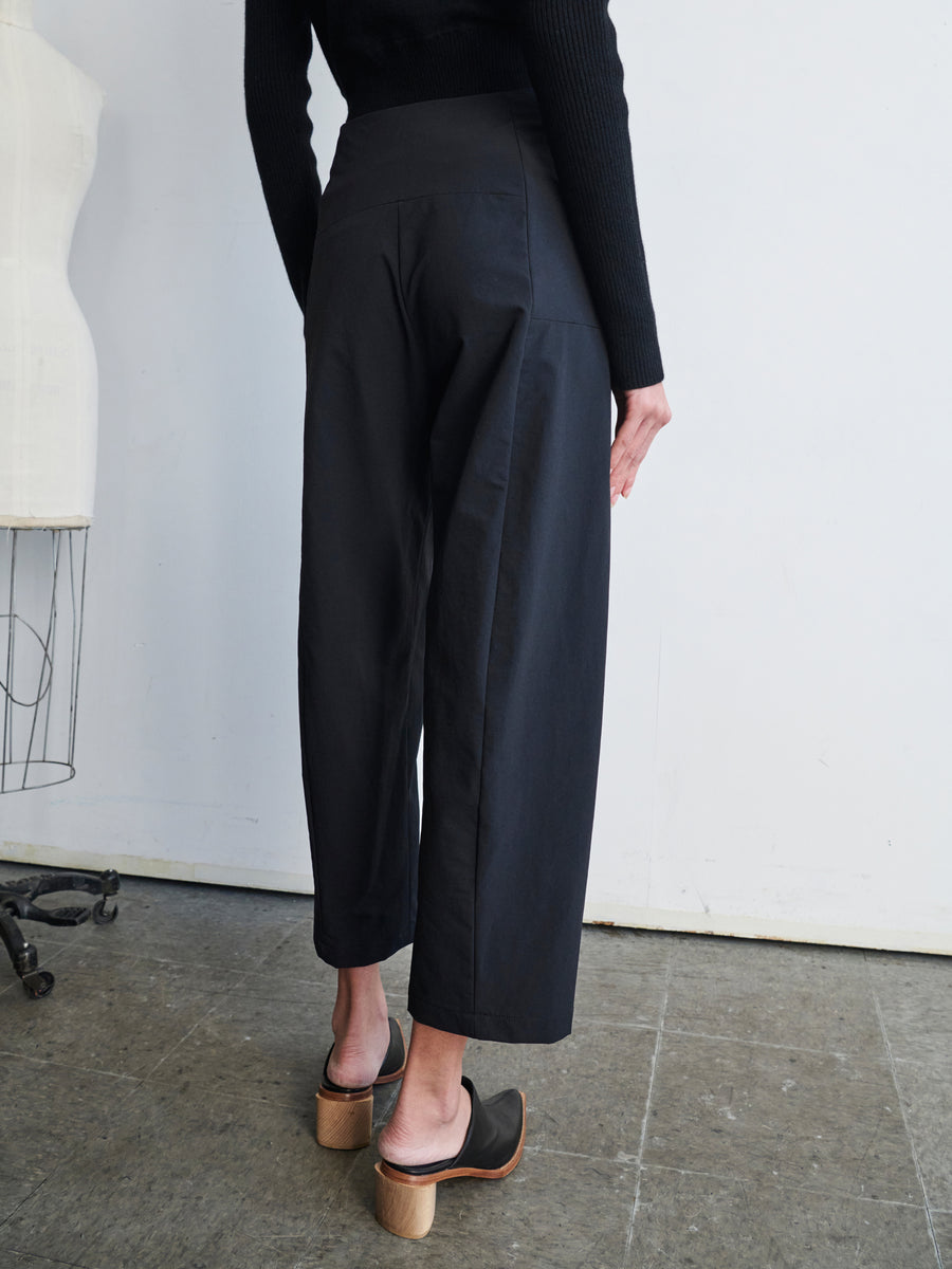 A person wearing the Margot Pant by Zero + Maria Cornejo and heeled clogs stands with their back to the camera. They are in a room with a dress form and a light-colored wall. The concrete floor adds an industrial touch to the scene.