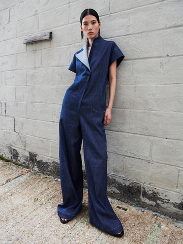 A model stands against a white brick wall wearing the Edi Boiler Suit by Zero + Maria Cornejo. The outfit, made from GOTS-certified cotton, features a high collar and diagonal fastening. She has dark hair pulled back and wears black open-toe heels, embodying eco-friendly fashion.