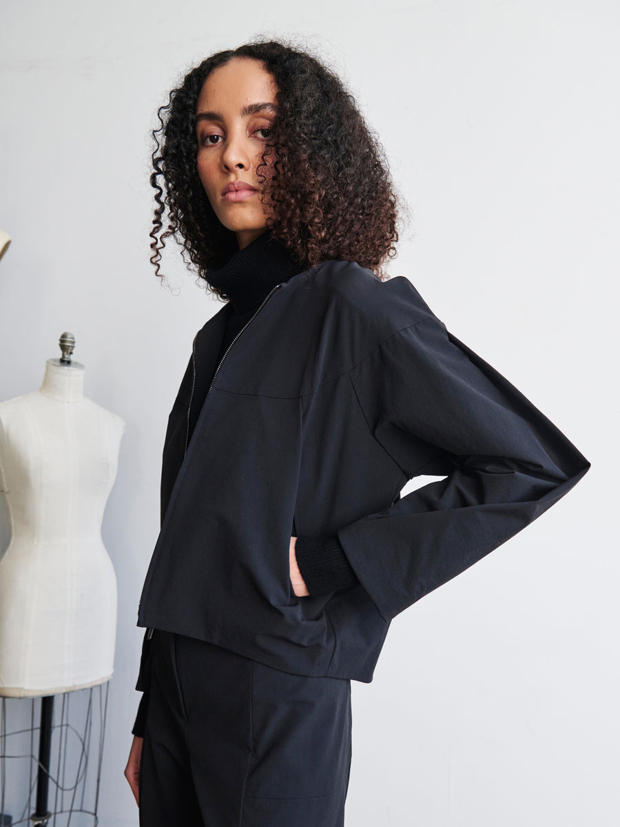 A person with curly hair stands confidently with one hand in their pocket, dressed in a stylish black outfit featuring a modern Ita Stretch design. In the background, a dress form mannequin showcases the T Bomber jacket by Zero + Maria Cornejo, complete with technical stretch material and a two-way front zipper.