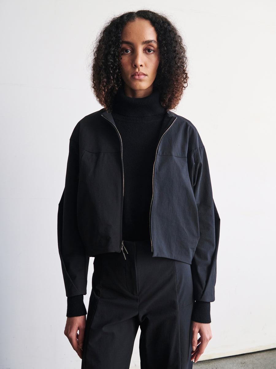 A person with curly hair stands against a plain white background, wearing a sleek modern T Bomber jacket from Zero + Maria Cornejo paired with a black turtleneck. The jacket features a stylish two-way front zipper. They have a calm expression, and their hands are out of view.