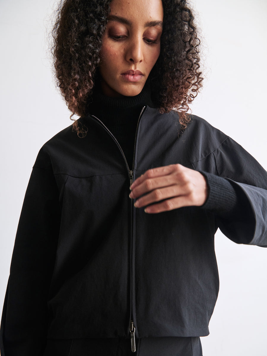 A person with curly hair is wearing the sleek black T Bomber jacket by Zero + Maria Cornejo, which features a two-way front zipper, over a black turtleneck. They are looking down, adjusting the cuff of their sleeve. The plain white wall in the background enhances the modern Ita Stretch vibe of the scene.