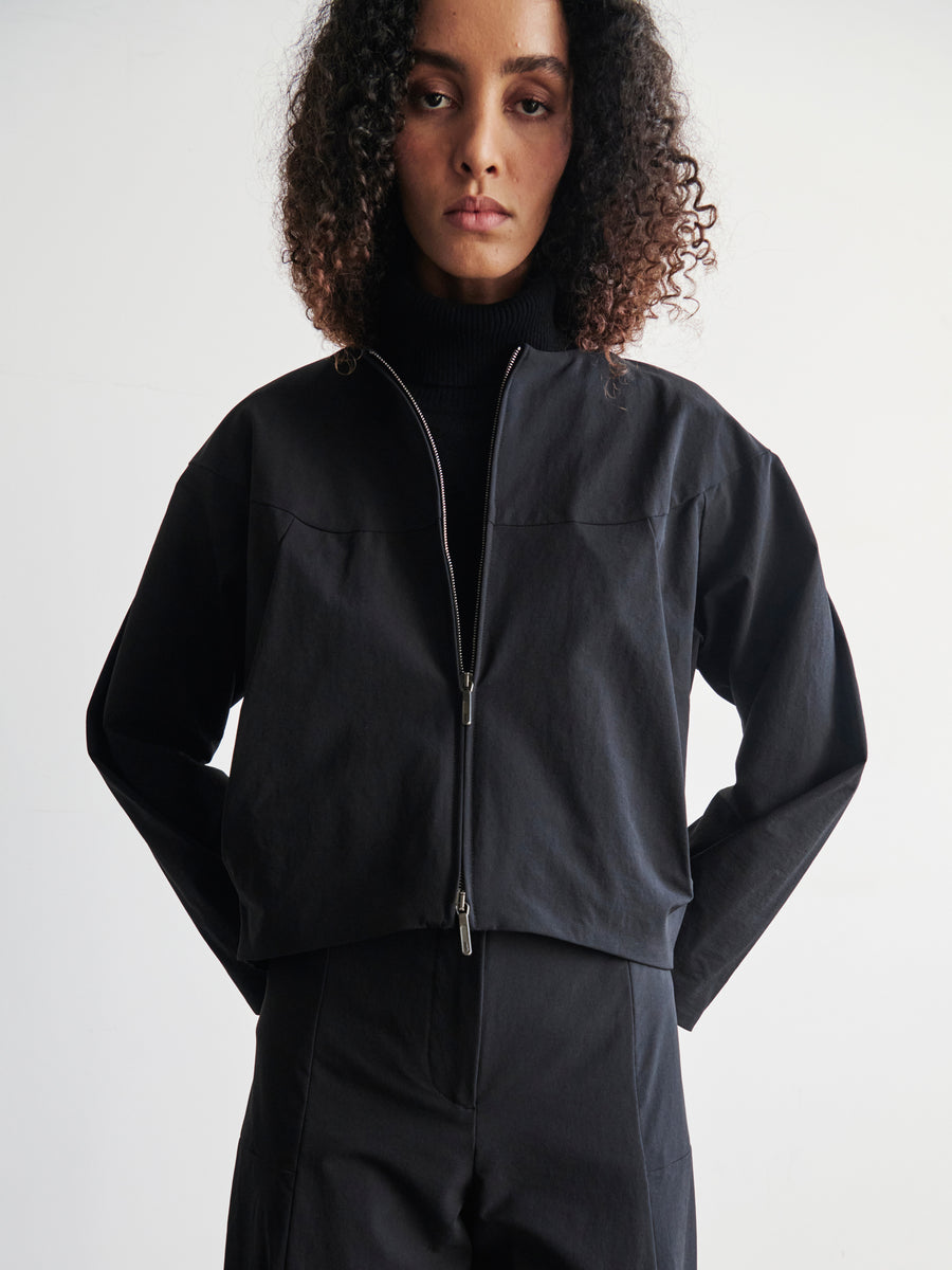 A person with curly hair is wearing a modern T Bomber by Zero + Maria Cornejo, featuring a sleek two-way front zipper, while standing against a plain white background with their hands behind their back.