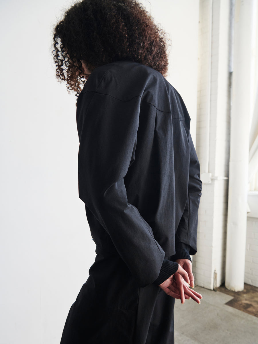 A person with curly hair stands in a minimalistic room, dressed in a modern T Bomber by Zero + Maria Cornejo. They face away from the camera with their hands clasped behind their back as natural light filters through a large window.