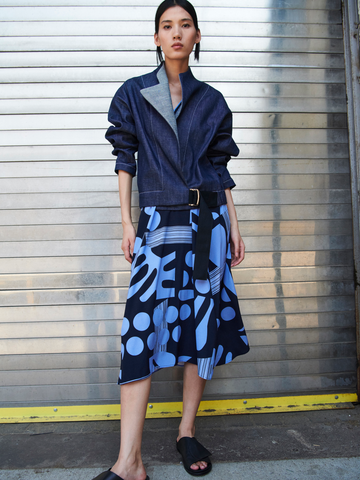A woman stands in front of a metal garage door, wearing a Zero + Maria Cornejo Long-Sleeved Edi Bomber with rolled-up sleeves over a blue skirt featuring abstract patterns. She has on black sandals and looks directly at the camera with a serious expression.