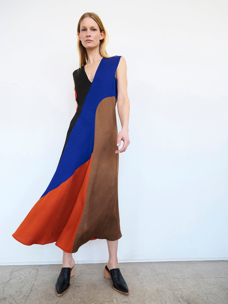 A person with long hair stands against a white background, wearing the Eve Mosa Dress by Zero + Maria Cornejo, which features an abstract color block design in blue, brown, orange, and black. They pair the sleeveless recycled drape dress with black heeled mules and pose with one foot slightly forward, enhancing the dynamic flow of this piece of eco-friendly fashion.