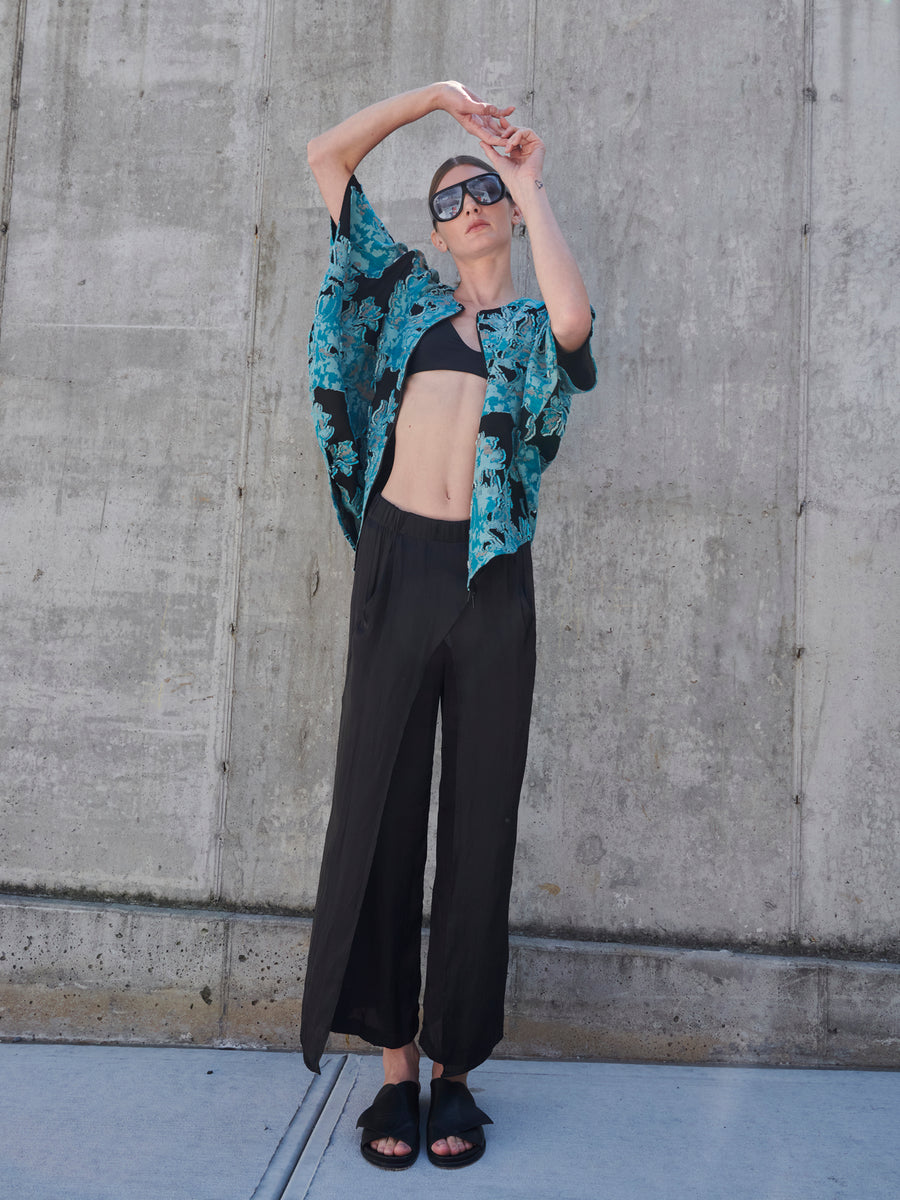 Wearing oversized sunglasses, a person poses casually against a concrete wall. They sport a blue patterned shirt over a black crop top, lightweight Ido Pants by Zero + Maria Cornejo, and sandals. Their arms are raised above their head in a carefree manner.