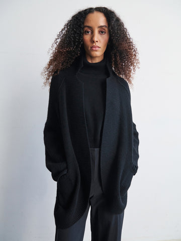 A person with long curly hair is wearing the Long-Sleeved Rio Shrug from Zero + Maria Cornejo, along with black pants. They stand against a plain white background with their hands in the shrug's pockets, maintaining a serious expression while looking directly at the camera.