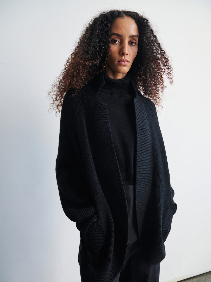 A person with long, curly hair is dressed in a black turtleneck paired with the oversized Long-Sleeved Rio Shrug from Zero + Maria Cornejo, crafted from recycled cashmere. They stand against a plain white background, looking directly at the camera.