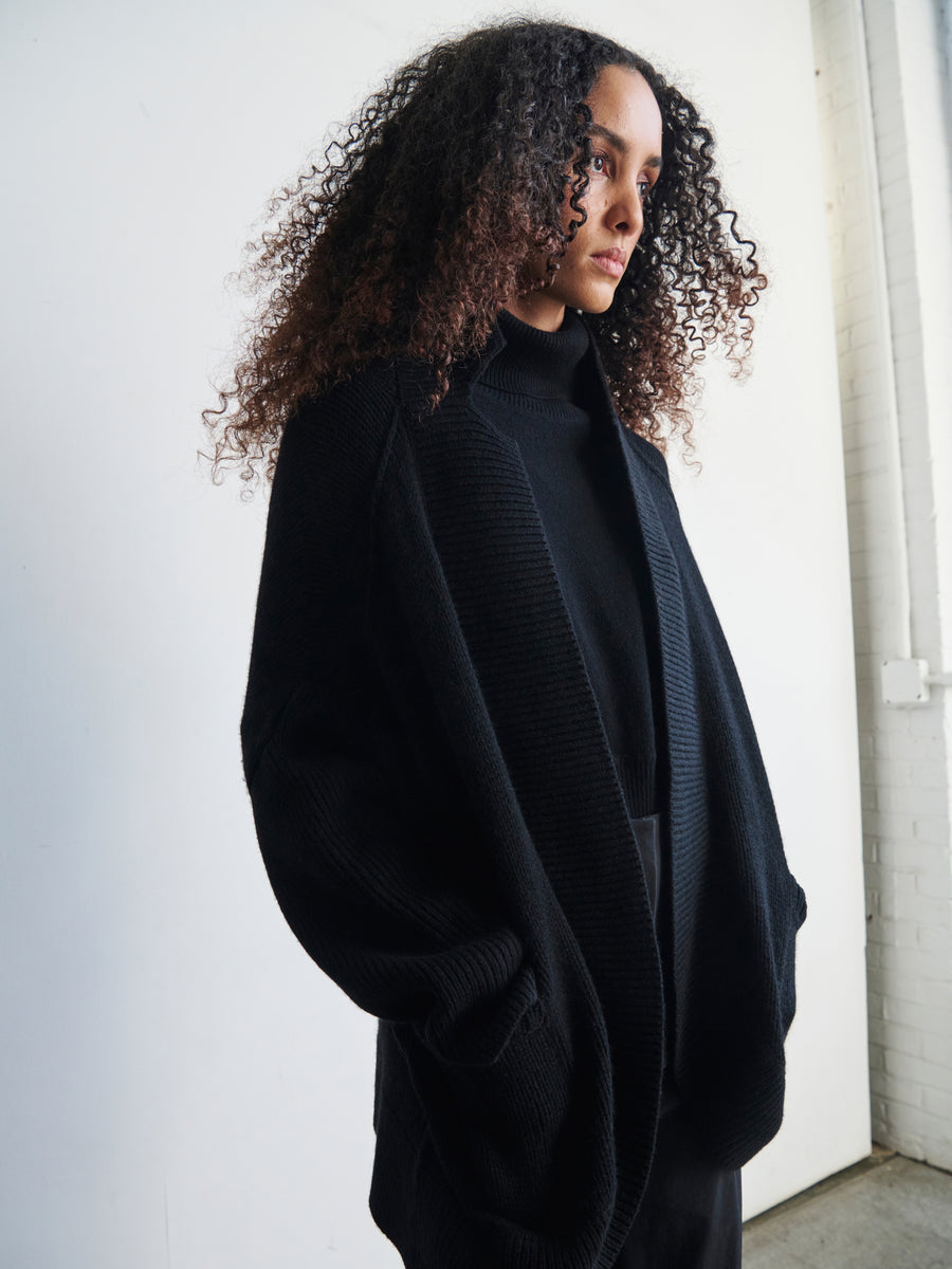 A woman with curly hair, wrapped in the Black Long-Sleeved Rio Shrug by Zero + Maria Cornejo, stands against a white background, gazing to the side. The lighting creates soft, natural shadows.