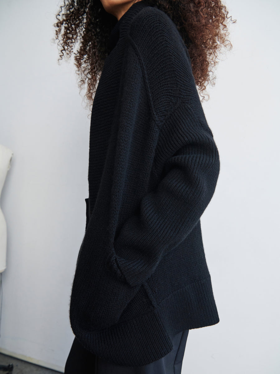 Against a neutral background, a person with curly hair is wearing the Long-Sleeved Rio Shrug by Zero + Maria Cornejo. This black oversized knit jacket, crafted from recycled cashmere, showcases a cozy textured design.