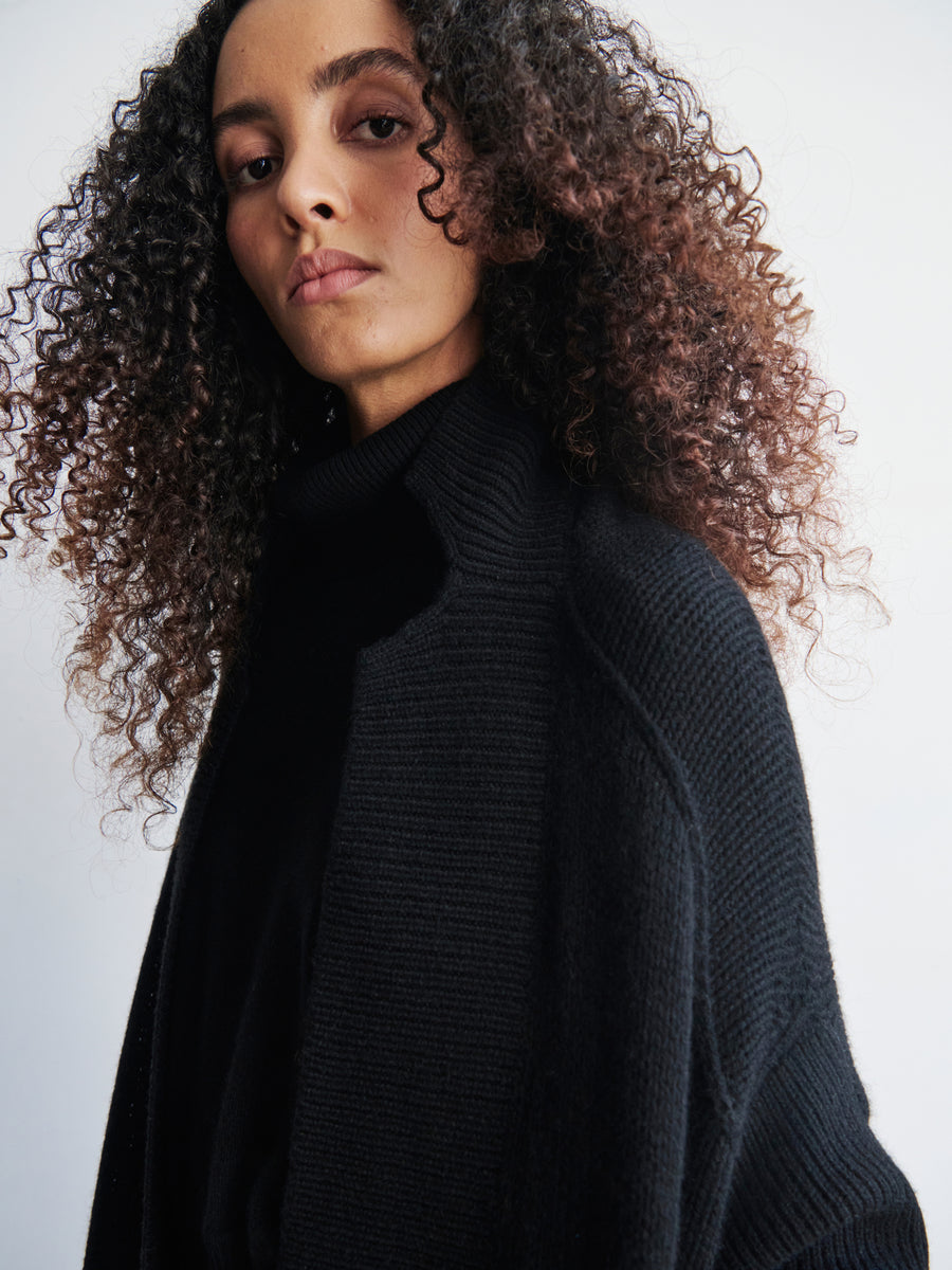 A person with curly hair, dressed in a black turtleneck sweater and the Long-Sleeved Rio Shrug by Zero + Maria Cornejo, is elegantly draped in a graceful silhouette while looking at the camera against a light background.