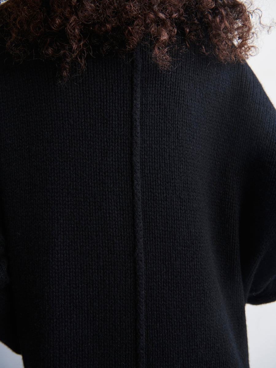 A person with curly hair is shown from the back, wearing the Long-Sleeved Rio Shrug by Zero + Maria Cornejo, a black textured knit jacket. The focus highlights the garment's recycled cashmere material and vertical seam, showcasing its elegantly draped silhouette.