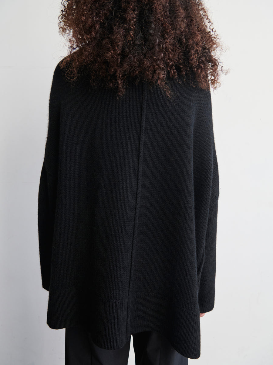 A person with curly hair is seen from the back wearing a loose-fitting Long-Sleeved Rio Shrug in black, designed by Zero + Maria Cornejo, made from recycled cashmere. The background features a plain white wall.