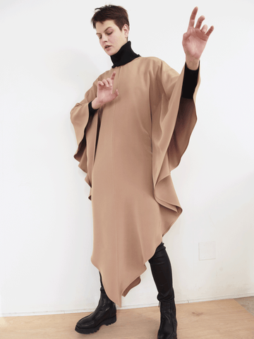 A person poses against a white wall in the fashionable Triangle Caftan by Zero + Maria Cornejo, crafted from responsible textiles. They wear it over a black turtleneck and pants with black boots, arms raised and fingers extended in an expressive stance.