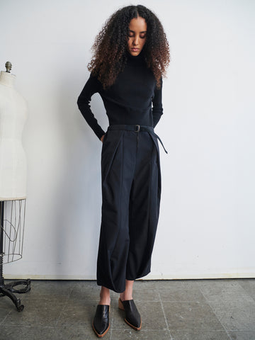 A person with curly hair stands in front of a plain white wall, wearing a black turtleneck and the Wrap Akeo Pant from Zero + Maria Cornejo. They have black slip-on shoes and are looking down. A dress form is partially visible on the left.