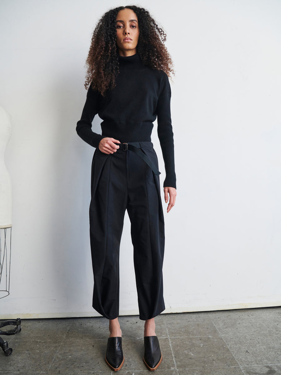 A person with curly hair stands against a white wall, dressed in a black turtleneck sweater and the high-waisted Wrap Akeo Pant made from technical-stretch woven fabric by Zero + Maria Cornejo, featuring an adjustable belt. Black pointed shoes complete the ensemble, while a dress form is partially visible on the left.