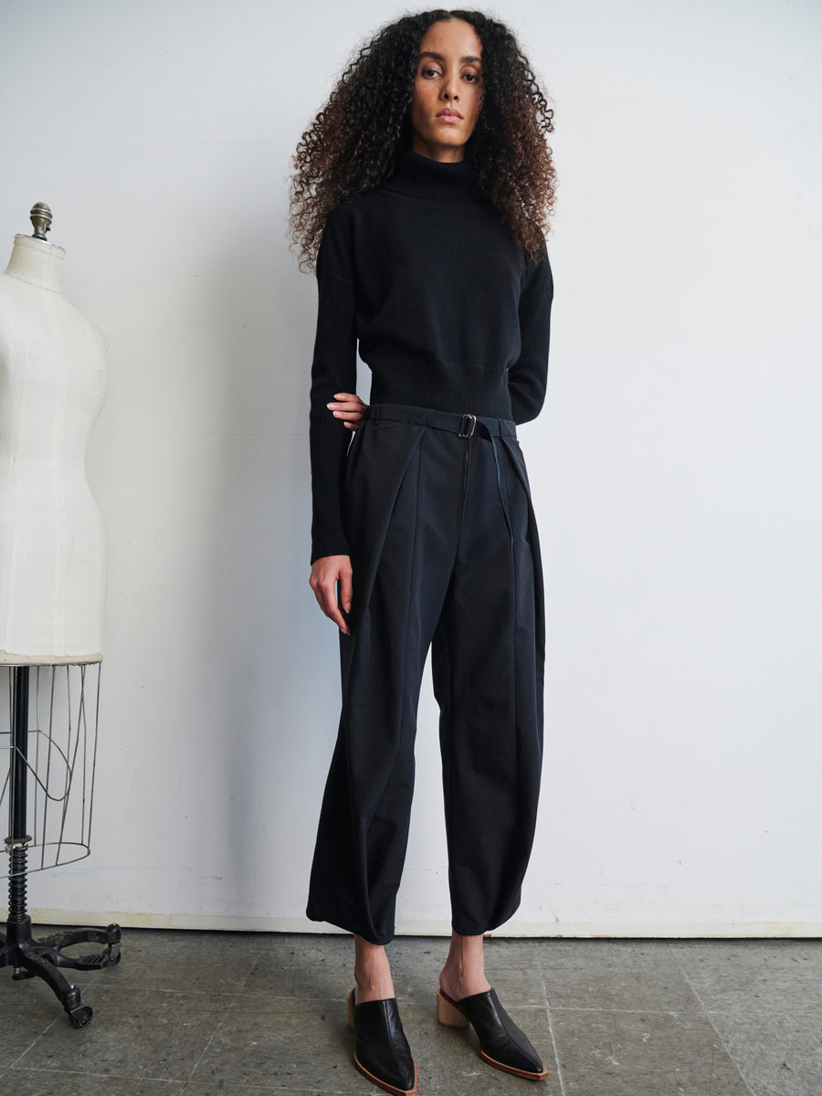 A woman with curly hair, dressed in a black turtleneck and the Wrap Akeo Pant by Zero + Maria Cornejo, stands in a minimalistic room next to a mannequin. She wears black slip-on shoes made from eco-hybrid fabric, with her hands relaxed by her sides.