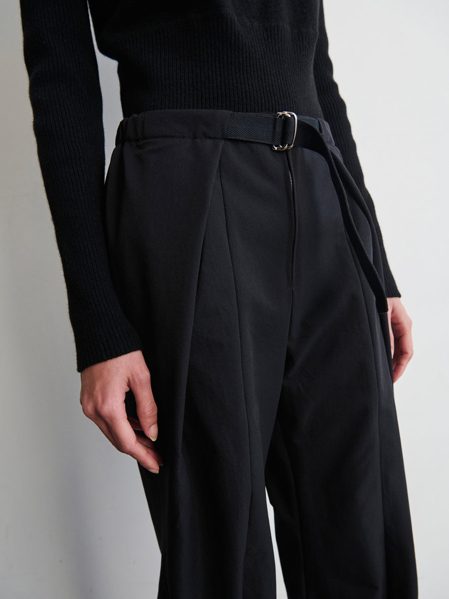 A person wears the Wrap Akeo Pant by Zero + Maria Cornejo, high-waisted black trousers made from wrinkle-resistant eco-hybrid fabric, featuring pleats and an adjustable belt, paired with a black long-sleeve sweater. Their hand rests by their side against a plain, light-colored background.