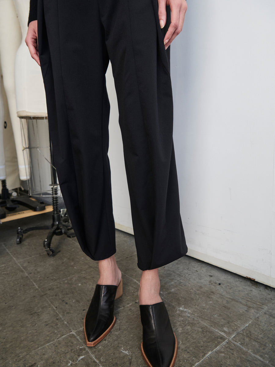 A person wearing the Wrap Akeo Pant by Zero + Maria Cornejo, crafted from wrinkle-resistant eco-hybrid fabric, and black slip-on shoes stands in a room with a concrete floor. Two mannequin legs are visible in the background to the left.